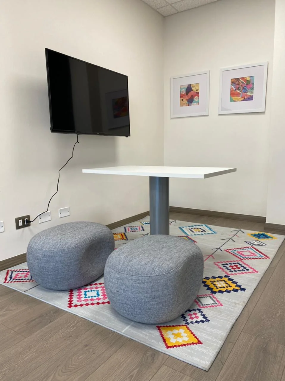 Espacio compartido con mesa, asientos grises, alfombra de colores y dos cuadros en la pared.