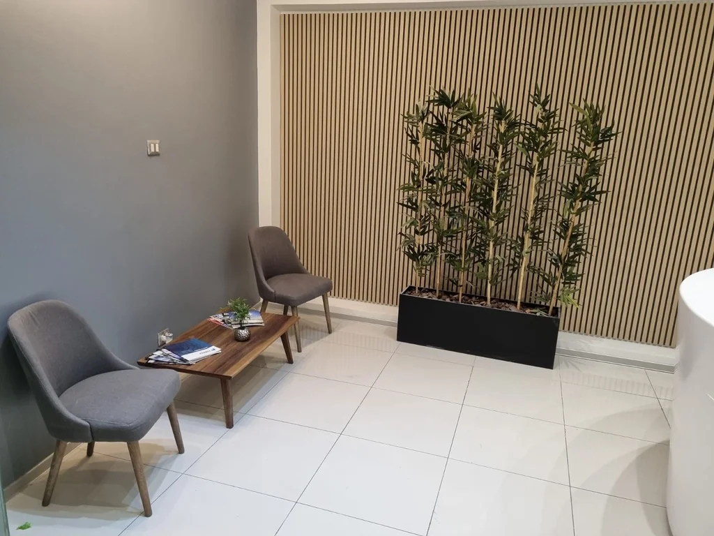 Zona de espera con asientos grises, mesa baja, plantas y revestimiento de listones de madera.